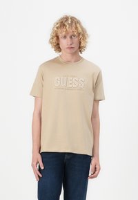 Beige bomull t-skjorte med rund hals og korte ermer, med hvit tekstgrafikk som leser "GUESS" og "CLASSIC AMERICAN STYLE."