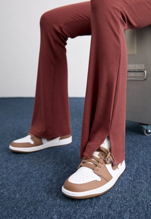 Beine, die rostfarbene Hosen mit seitlichen Schlitzen tragen, kombiniert mit weißen und braunen Sneakers auf blauem Teppich, sitzend auf grauen Möbeln.