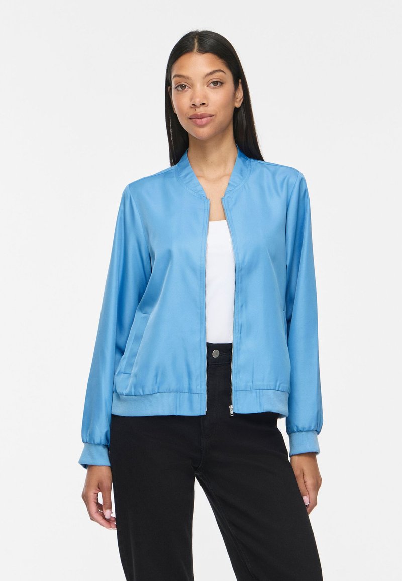 Femme portant une veste bomber bleu clair zippée sur un haut blanc et un pantalon noir, debout devant un fond blanc uni.