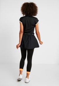 Kvinna med lockigt hår står vänd bort, iförd en svart kortärmad topp, svart kjol, svarta leggings med orange muddar och vita sneakers.