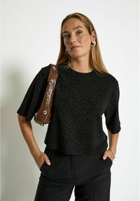 Juodas tekstūruotas trumpas rankovėmis marškinėliai su banginiu raštu. Modelis laiko rudos spalvos rankinę su sidabro akcentais ir nešioja sidabro apyrankes.