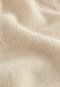 Cremefarbener, gestrickter Stoff mit einer strukturierten, weichen Oberfläche, die ein sichtbares Nähmuster und leichte Variationen in der Dicke aufweist.