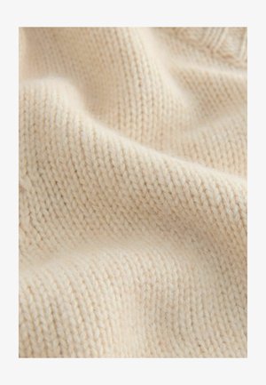 Tela de punto de color crema que muestra una superficie texturada y suave con un patrón de costura visible y ligeras variaciones en el grosor.