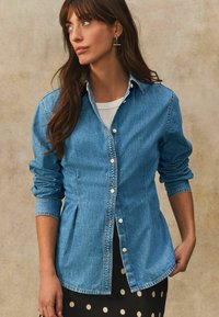 Lichte blauwe denimhemd met knoopsluiting aan de voorkant, lange mouwen en een afgeronde zoom. Draag het over een witte blouse en een zwarte rok met stippen.