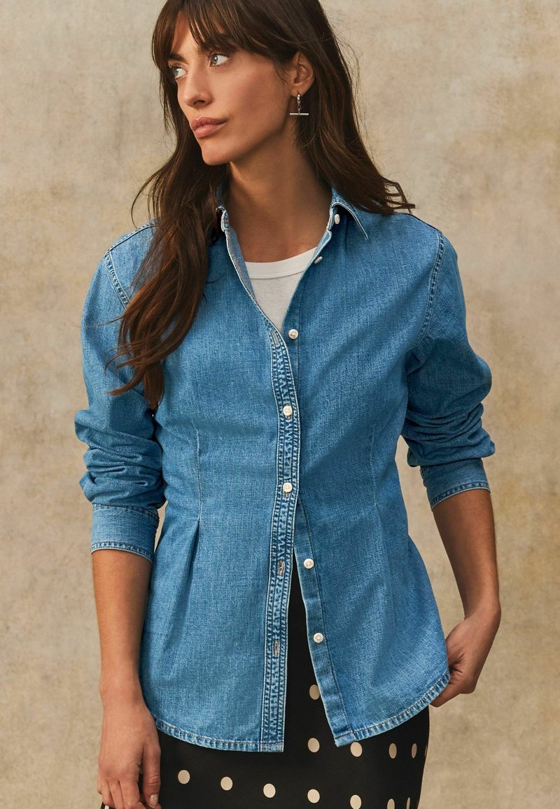 Lichte blauwe denimhemd met knoopsluiting aan de voorkant, lange mouwen en een afgeronde zoom. Draag het over een witte blouse en een zwarte rok met stippen.