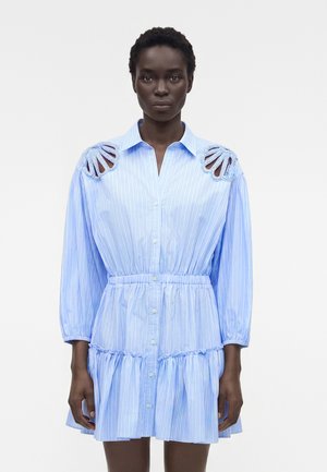 Γυναίκα που φοράει φωτεινό γαλάζιο ριγέ φόρεμα με κουμπιά μπροστά, σούρα στη μέση, βολάν στο τελείωμα και διακοσμητικές εγκοπές στους ώμους.