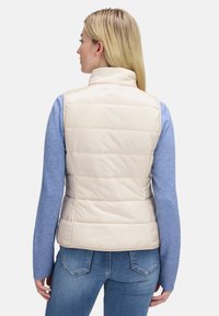 Puchowy kamizelka z wysokim kołnierzem, pikowanym wzorem i beżowymi wykończeniami, noszona na jasnoniebieskim swetrze, zestawiona z niebieskimi dżinsami.