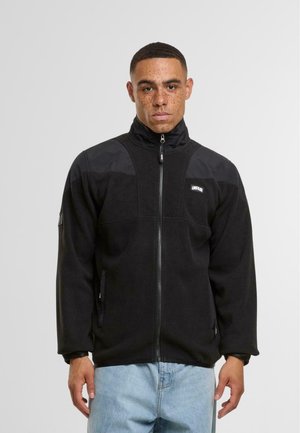 Giacca in pile nera con colletto alto, chiusura a zip e pannelli in nylon nero a contrasto sulle spalle. Presenta tasche laterali e un'etichetta con logo.