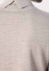 Gros plan sur une personne portant un pull tricoté beige clair avec un col plié, mettant en valeur le tissu texturé et la zone de l'épaule.