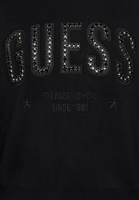Sweat-shirt noir avec logo "GUESS" en relief orné de strass noirs. Présente un texte plus petit en dessous indiquant "LOS ANGELES-CAL SINCE 1981."