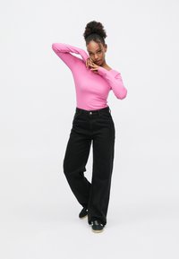 Heleroosa soonikkoega pikkade varrukatega top, mida kannab mustad kõrge tulbiga laiade säärtega teksapüksid. Modell seisab neutraalsel taustal, et näidata jalatsite detaile.