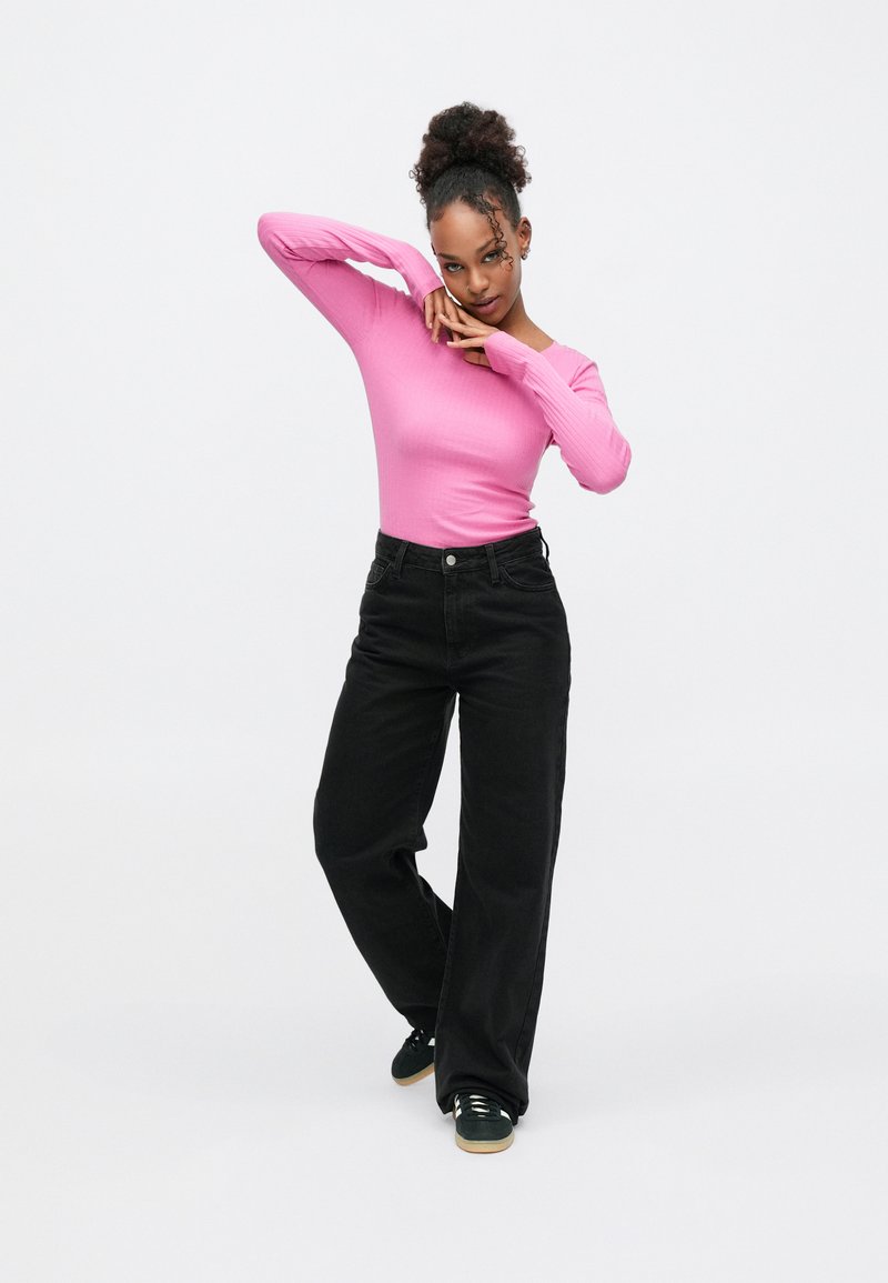 Hellrosa geripptes Langarmshirt kombiniert mit schwarzen, hoch taillierten Jeans mit weitem Bein. Das Model steht vor einem neutralen Hintergrund und zeigt Details der Schuhmode.