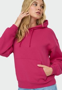 Jeune femme aux longs cheveux blonds portant un sweat à capuche rose vif avec une poche avant, posant avec une main près de son visage et l'autre dans sa poche.