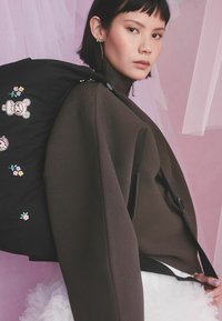 Noor naine lühikeste tumedate juustega, pruunis jopes, kandmas musta seljakotti, millel on lilleline ja karukujuline tikand roosal taustal.