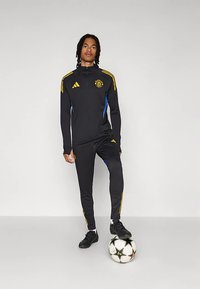 Juodas sportinis kostiumas su geltonais ir mėlynais akcentais, su užtrauktuku viršuje ir derančiomis kelnėmis. Modelis stovi ant juodai baltos futbolo kamuolio.