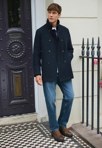 Marineblauer doppelt geknöpfter Mantel, weißes T-Shirt, blaue Jeans und braune Schnürstiefel. Steht neben einer dekorativen Tür mit kunstvollen Details.