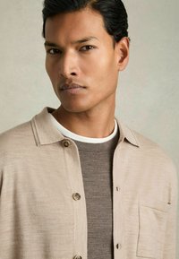 Camisa de botón beige claro con cuello corto, que presenta dos bolsillos frontales y una textura sutil, superpuesta sobre una camiseta ajustada de color marrón oscuro.
