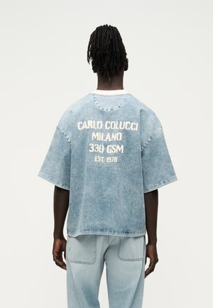 Άνδρας φοράει ανοιχτό μπλε, φαρδιά τζιν πουκάμισο με το κείμενο «Carlo Colucci Milano 330 GSM Est. 1978» στην πλάτη, συνδυασμένο με ανοιχτό μπλε τζιν παντελόνι.