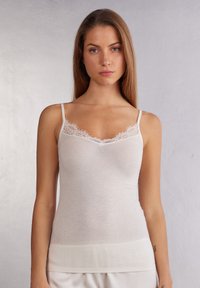 Camisole beige clair avec des fines bretelles, ornée d'une dentelle le long de décolleté. Fabriquée en matériau doux, avec une texture lisse.