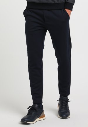 Homme portant un pantalon noir ajusté et des baskets noires avec des accents beige, debout les mains dans les poches devant un fond uni.