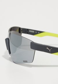 Lunettes de sport avec des verres teintés gris, monture grise et accents jaune fluo. Présentent un logo Puma sur la branche et des prises latérales en caoutchouc.