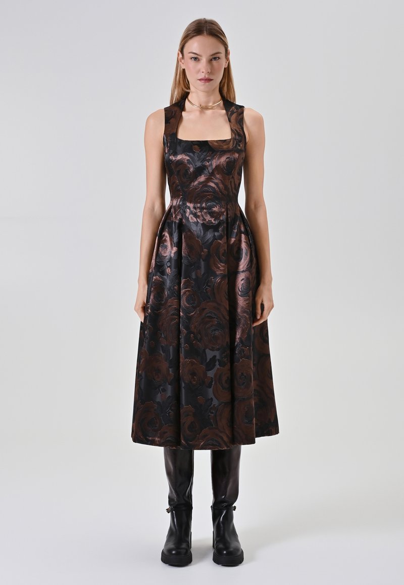 Schwarzes Blumenkleid mit quadratischem Ausschnitt und ausgestelltem Rock, verziert mit bronzefarbenen Rosenmustern und transparenten Trägern. Kombiniert mit schwarzen Ankle Boots.