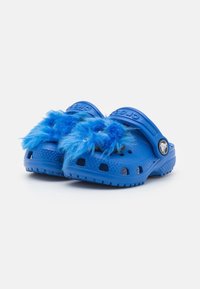 Modré Crocs s chlupatou modrou kožešinou na vrchu, s pásky na patě a ventilačními otvory na bílém pozadí.