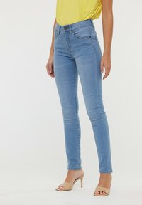 Jeans skinny bleu clair en denim, avec une taille mi-haute, cinq poches et un léger effet délavé. Portés avec des talons ouverts beiges.