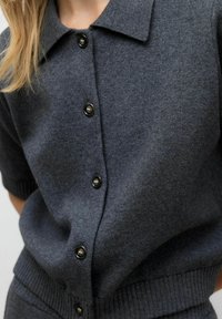 Grå stickad topp med klassisk krage, korta ärmar och en rad svarta knappar framtill. Texturen är mjuk och slät.