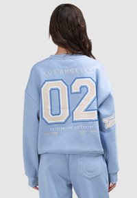 Sweat-shirt bleu clair avec un grand graphique "02", texte "LOS ANGELES" et détails brodés. Tissu doux et coupe décontractée.