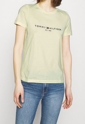 Vrouw draagt een lichtgroen Tommy Hilfiger T-shirt met logo en blauwe jeans, staand tegen een effen lichte achtergrond.