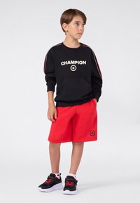 Ragazzo che indossa un felpa nera Champion, pantaloni corti rossi e scarpe da ginnastica nere con lacci rossi, in piedi con le mani nelle tasche e che guarda a destra.