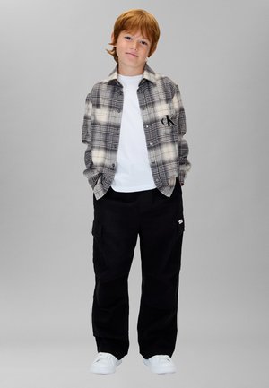 Graues und schwarzes Kariertes Hemd mit Knopfleiste und CK-Logo, darunter weißes T-Shirt, schwarze Cargohose und weiße Sneakers.