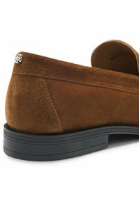 Καφέ σουέτ παπούτσι χωρίς κορδόνια με μαύρη καουτσούκ σόλα. Διαθέτει μια ασημένια διακοσμητική λεπτομέρεια στη φτέρνα και μια λεία, υφή επιφάνεια.