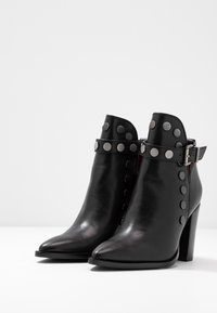 Černé kožené kotníkové boty s špičatou špičkou, zdobené stříbrnými kovovými nýty a páskem s přezkou kolem kotníku. Design s blokovým podpatkem.
