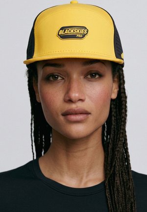 Woman with braided hair wearing a yellow and black cap labeled "BLACKSKIES PRO" and a black shirt, facing forward with a neutral expression.