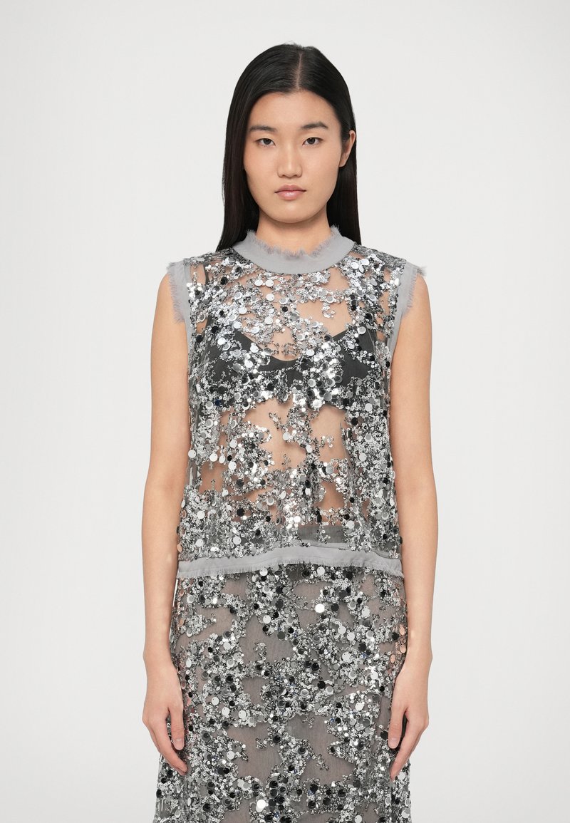 Διάφανο, αμάνικο τοπ με ασημένιες πούλιες, γκρι ξεφτισμένο γιακά και λεπτή υφή υφάσματος. Διαφανή στρώματα δημιουργούν βάθος.