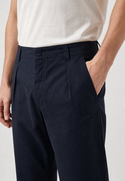 Homme portant un pantalon bleu foncé et une chemise blanche, la main droite dans la poche, sur un fond uni.