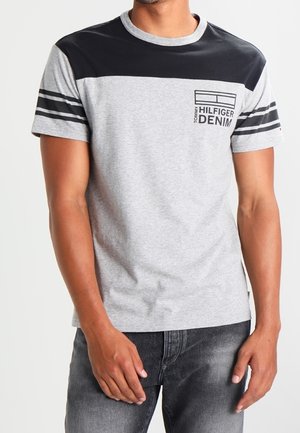 Mann trägt ein grau-schwarzes T-Shirt mit kurzen Ärmeln mit Tommy Hilfiger Denim-Logo und dunkelgraue Jeans, steht vor weißem Hintergrund.