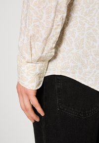 Chemise beige à manches longues avec un léger motif de feuilles et poignets boutonnés, portée avec un jean noir en denim, montrant une main détendue sur le côté.