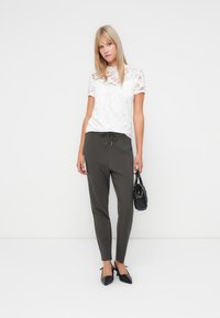 Hvit bluse i blonder med korte ermer, sammen med mørkegrå, smale bukser, svarte leiligheter og en liten svart håndveske. Enkelt og rent design.