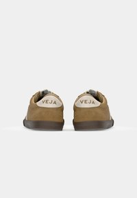 Καφέ σουέτ αθλητικά παπούτσια με λευκές λεπτομέρειες και υφασμάτινη σόλα. Λογότυπο "VEJA" στη φτέρνα, απλός σχεδιασμός, χαμηλή σιλουέτα.