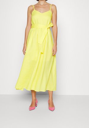 Geel mouwloze midi-jurk met dunne bandjes en een ceintuur in de taille, gecombineerd met roze pumps met een puntige neus op een effen achtergrond.