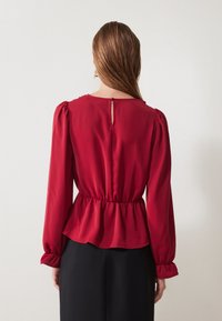 Červená blúzka s dlhým rukávom, ktorá má peplum dizajn, zberaný pás a kľúčovú dieru na chrbte. Hladká látka s jemným leskom.