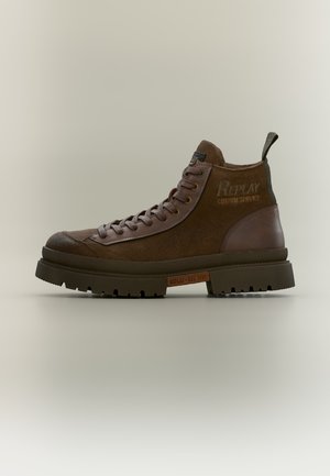 Botte montante en suède et cuir marron avec semelle épaisse en caoutchouc et lacets, arborant la marque "Replay" sur le côté et la semelle.