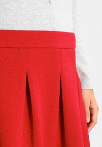 Gonna rossa con texture e motivo floreale, caratterizzata da pieghe e una cintura larga, abbinata a un maglione grigio chiaro con bottoni.