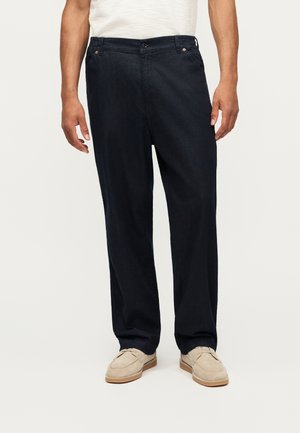 Homme portant un pantalon ample bleu marine foncé avec des mocassins en daim beige et une chemise blanche, debout devant un fond clair uni.