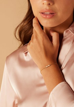 Bracelet en or avec trois accents en pierres blanches, porté sur une main légèrement bronzée. L'arrière-plan présente une teinte douce et neutre.