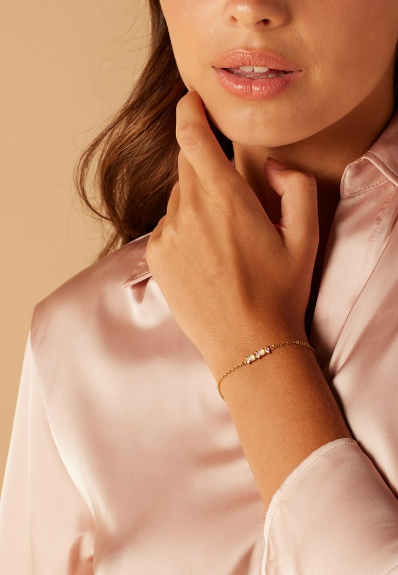 Gouden armband met drie witte steenaccenten, gedragen op een licht gebruinde hand. De achtergrond heeft een zachte, neutrale tint.