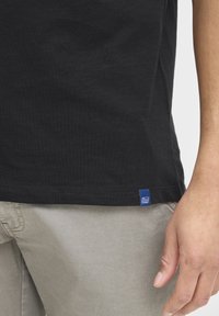 Crna pamučna majica s okruglim izrezom, s plavim detaljem oznake na rubu. Nosena s svijetlo sivoj hlačama, što daje ležeran izgled.